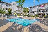 Located at 12200 Valley View St in Garden Grove, California, this outdoor courtyard space showcases a beautifully designed communal pool area that serves as the centerpiece of the property. The architecture follows a classic Spanish Colonial Revival style, characterized by white stucco exteriors, terracotta-tiled rooflines, arched entryways, and wrought iron balcony railings that frame the surrounding multi-story residential buildings.

The sparkling rectangular pool is finished with deep blue tile accents and is complemented by a generous surrounding deck that offers ample space for lounging and recreation. Sun loungers and seating areas are thoughtfully arranged poolside, providing residents with comfortable spots to relax under the Southern California sun. A covered pergola structure with diagonal lattice detailing offers shaded outdoor dining, while multiple patio tables with umbrellas are distributed across the courtyard for additional gathering options.

Tall, swaying palm trees and mature shade trees are interspersed throughout the courtyard, enhancing the tropical, resort-like ambiance. Lush landscaping with hedges and potted plants adds greenery and privacy throughout the space. The entire area is enclosed by decorative iron fencing, creating a secure and inviting environment. Under clear blue skies, this well-maintained courtyard captures the essence of relaxed Southern California outdoor living.