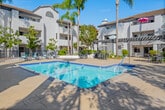 Located at 12200 Valley View St in Garden Grove, California, this outdoor courtyard space showcases a beautifully designed communal pool area that serves as the centerpiece of the property. The architecture follows a classic Spanish Colonial Revival style, characterized by white stucco exteriors, terracotta-tiled rooflines, arched entryways, and wrought iron balcony railings that frame the surrounding multi-story residential buildings.

The sparkling rectangular pool is finished with deep blue tile accents and is complemented by a generous surrounding deck that offers ample space for lounging and recreation. Sun loungers and seating areas are thoughtfully arranged poolside, providing residents with comfortable spots to relax under the Southern California sun. A covered pergola structure with diagonal lattice detailing offers shaded outdoor dining, while multiple patio tables with umbrellas are distributed across the courtyard for additional gathering options.

Tall, swaying palm trees and mature shade trees are interspersed throughout the courtyard, enhancing the tropical, resort-like ambiance. Lush landscaping with hedges and potted plants adds greenery and privacy throughout the space. The entire area is enclosed by decorative iron fencing, creating a secure and inviting environment. Under clear blue skies, this well-maintained courtyard captures the essence of relaxed Southern California outdoor living.