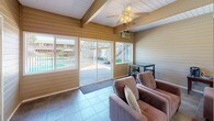 This inviting single-floor space showcases a relaxed, casual aesthetic rooted in warm architectural details. Horizontal wood-paneled walls in a soft golden tone run throughout, complemented by white-painted vaulted ceilings with exposed wooden beams that add both height and character to the room. Ceiling fans are positioned throughout to ensure comfortable airflow.

The central gathering area is generously sized, anchored by a classic brick fireplace framed by large windows that flood the space with natural light. Multiple seating arrangements — including plush sofas, loveseats, and armchairs — are thoughtfully arranged to encourage conversation and relaxation. A dark wood coffee table and side tables accent the seating areas, while decorative touches such as framed artwork, lamps, and potted greenery add warmth and life to the interior.

Stone tile flooring in a neutral, earthy tone runs throughout the space, lending durability and a grounded aesthetic that ties the room together. A dining or bar-height table with stools is positioned near the windows, providing a casual spot for small gatherings.

Large sliding glass doors open toward an outdoor pool area, connecting the interior to the exterior and allowing for an abundance of natural light and open views. The overall ambiance is cozy yet spacious, making this a welcoming communal retreat.