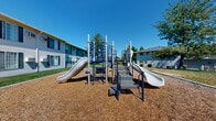 This multi-story residential community spans three floors and is thoughtfully designed to foster a welcoming, communal atmosphere. The exterior of the buildings features classic white siding accented with navy blue shutters and a neutral gray roofline, creating a clean and cohesive aesthetic across the property.

The outdoor spaces are a standout feature of this community. A centrally located playground is equipped with multiple slides, climbing structures, and blue metal framework, set on a wood chip safety surface — ideal for younger residents. Adjacent to the playground, a poured concrete patio provides a dedicated area for outdoor gatherings, complete with picnic tables and a charcoal grill for residents to enjoy.

Lush, manicured green lawns surround the buildings, dotted with mature deciduous and evergreen trees that offer natural shade and privacy. Ornamental shrubs with seasonal color add visual interest to the building entrances, enhancing the overall curb appeal. Wide concrete walkways connect various areas of the property, making navigation throughout the community easy and accessible.

The grounds are open and spacious, with a clear sense of room between buildings, allowing for natural light and open sightlines. The bright, sunny setting underscores the community's welcoming outdoor character, making this a well-rounded residential property designed for comfortable, connected living.