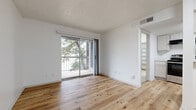 This single-floor residence is designed with a clean, modern aesthetic, characterized by crisp white walls, textured ceilings, and light wood-look flooring that flows throughout the main living areas. The living room is a standout space, bathed in natural light streaming through a large sliding glass door that opens directly onto a private balcony. The balcony is enclosed by a metal railing and overlooks mature trees, providing a serene, leafy backdrop. An adjacent area features built-in wall shelving, offering practical display and storage solutions within the open layout.

The kitchen is efficiently laid out and equipped with stainless steel appliances, including an electric range with overhead ventilation and a full-size refrigerator. White cabinetry and light countertops contribute to the kitchen's clean, contemporary finish. The living and kitchen areas connect openly, enhancing the sense of spaciousness throughout.

The bedroom is carpeted for comfort and features a ceiling fan with a light fixture, a window with blinds offering outdoor views, and access to a closet. Overall, the unit presents a well-balanced combination of functional design and comfortable, light-filled living spaces.