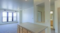 This single-floor space offers a clean, open-concept layout that seamlessly connects the kitchen and living areas. The kitchen features light wood cabinetry with ample storage, a neutral-toned countertop, and a generous peninsula that serves as both a workspace and a casual dining or gathering surface. A distinctive pass-through window cut into the wall bridges the kitchen to an adjacent room, enhancing the sense of openness and connectivity throughout the space.

The living area is finished with wall-to-wall carpeting that adds warmth underfoot, while crisp white walls keep the atmosphere bright and neutral. Track lighting mounted on the ceiling provides versatile, adjustable illumination across the main living space. A large window along one wall invites an abundance of natural light, contributing to the airy feel of the interior.

A set of doors near the window area suggests access to closets or additional storage, adding to the unit's practical layout. The overall design is straightforward and functional, with a light, neutral palette throughout that offers a versatile canvas for personalization. The space is well-suited for those seeking a bright, connected living environment with thoughtful built-in features.