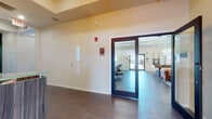 Located at 73 Brownstone Way in Englewood, NJ, this single-floor community amenity space is thoughtfully designed to offer residents a polished and functional gathering environment. The entrance features a sleek reception desk with a textured wood-panel facade, set beneath a striking geometric chandelier with Edison-style bulbs, complemented by a bold green accent wall and wall-mounted television — establishing an inviting first impression.

The main lounge area is expansive and open, anchored by rich, dark hardwood flooring throughout. A cozy seating arrangement with a leather sofa and accent chairs surrounds a classic white fireplace topped with a wall-mounted TV, framed by the deep green feature wall. Large windows allow ample natural light to fill the space, creating a warm and airy ambiance.

Adjacent to the lounge, a game zone includes a full-size pool table illuminated by elegant pendant lighting, alongside additional casual seating. A wet bar and kitchenette area with bar-height counter seating provides a convenient spot for refreshments and casual gatherings.

Textured chevron-patterned accent walls add visual depth in key areas, while recessed lighting and tray ceilings contribute to the refined aesthetic. Double glass doors lead to outdoor areas, maintaining a seamless connection between the interior and the surrounding environment.