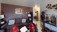 This single-floor residence offers a cohesive and well-appointed layout across its main living spaces. The living room makes a strong first impression with its bold, deep burgundy accent wall, textured ceilings, and tile flooring. It is furnished with a comfortable seating arrangement, decorative vases, and warm artwork, creating an inviting and personalized atmosphere.

The kitchen features rich wood cabinetry extending to the ceiling for ample storage, dark granite-style countertops, a stainless steel sink, and black appliances including an electric range and refrigerator. The tile flooring continues seamlessly from the living area into the kitchen.

The two bedrooms are carpeted and offer a cozy, quiet retreat. Each room is comfortably sized to accommodate a queen bed with nightstands and additional furniture. One bedroom features a set of louvered bifold closet doors providing generous storage, while both rooms display thoughtful décor accents. Soft, curtained windows allow for controlled natural light.

The bathroom includes a single-sink vanity with dark countertop and wood cabinetry, tile flooring, a full bathtub with a shower curtain, and practical over-toilet shelving for additional storage. Wall-mounted décor and organizational accessories add functional character to the space throughout.
