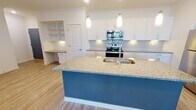 This well-appointed single-floor residence offers a bright, open-concept layout that seamlessly connects the kitchen, dining, and living areas. The kitchen is a standout feature, showcasing crisp white shaker cabinets, granite countertops, and a striking blue-accented central island with an undermount sink and pendant lighting overhead. Stainless steel appliances — including a refrigerator, range, and microwave — complement the subway tile backsplash and under-cabinet lighting, creating a clean, modern aesthetic. A built-in shelving nook and a dedicated desk area add functional charm near the kitchen. Light wood-style flooring flows throughout the main living spaces, while the living area is equipped with a ceiling fan and access to natural light through a window. The bedrooms are carpeted and generously sized, with large windows that bring in abundant natural light and offer neighborhood views. The unit includes two bathrooms: one featuring a tub/shower combination with tile surround and granite vanity, and another with a walk-in shower, white tile surround, and a matching granite-topped vanity. Throughout the space, a neutral white color palette, crown molding details, and consistent finishes create a cohesive and inviting atmosphere.