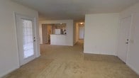 This single-story residence offers an open-concept floor plan that connects the main living area to the kitchen, creating a spacious and functional layout. The living room features neutral-toned carpet and is filled with natural light from a glass-panel door, lending the space a bright and airy feel. A smooth transition from carpet to tile flooring marks the boundary between the living and kitchen areas.

The kitchen is designed with light wood cabinetry, a white refrigerator, and a peninsula-style breakfast bar that serves as a natural divider between the cooking and living spaces. Recessed lighting illuminates the kitchen area, adding warmth to the overall neutral color palette of white and beige tones throughout.

A hallway visible from the main living space leads to additional rooms, suggesting a practical and well-organized floor plan. The walls are finished in a clean, off-white tone that complements the light flooring and cabinetry, contributing to a cohesive and understated aesthetic. Overall, the space presents a functional, move-in-ready layout with a comfortable and straightforward design.