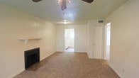 This single-floor space centers around a carpeted living room that provides a comfortable, open feel. A fireplace with a simple mantel serves as a focal point along one wall, adding a classic touch to the room. Overhead, a ceiling fan with a light fixture ensures year-round comfort and adequate illumination. The textured ceiling adds subtle visual interest to the space.

The living room connects directly to what appears to be a kitchen or utility area, visible through a doorway that allows natural light to filter into the main space. White cabinetry can be seen in the adjacent room, suggesting a functional kitchen layout. A second door provides access to an additional room, contributing to the overall flow and connectivity of the floor plan.

Neutral-toned walls throughout maintain a clean, understated aesthetic, while the carpeted flooring adds warmth underfoot. A wall-mounted vent indicates a central HVAC system for climate control. The layout is practical and well-organized, making efficient use of the available space while maintaining clear transitions between areas.