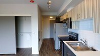 This single-floor space showcases a contemporary design with a cohesive, modern aesthetic throughout. The kitchen is a standout feature, outfitted with stainless steel appliances including a refrigerator, microwave, and electric range, complemented by light countertops and two-toned cabinetry in dark and textured finishes. A stainless steel sink adds a polished touch, while track lighting overhead ensures the space is well-lit and functional. The dark hardwood flooring runs consistently throughout, grounding the space with warmth and contrast against the light-colored walls and ceilings. A galley-style layout keeps the kitchen efficient and organized, with ample upper cabinet storage. Adjacent to the kitchen, a hallway leads toward the entry door, maintaining a clean sightline through the unit. A walk-in closet area is visible off the main corridor, featuring built-in shelving and hanging rods, offering generous storage solutions. The overall ambiance is clean, modern, and practical, with neutral tones and quality finishes that create a well-appointed living environment suitable for everyday comfort.