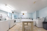 Located at 2441 Finch Avenue West in Toronto, this shared laundry facility is a well-organized, functional space spread across a single floor. The main laundry room is finished with light blue walls and classic cream-toned floor tiles, creating a clean and bright environment illuminated by fluorescent overhead lighting. The room accommodates multiple coin-operated washing machines and dryers arranged along the walls, offering ample capacity for residents. A central folding table provides a convenient surface for sorting and folding laundry. A utility sink along one wall is paired with a tenant information bulletin board nearby, keeping residents informed of building notices and guidelines. A cast-iron radiator ensures comfortable temperatures year-round. The facility also includes a secondary room accessible through an interior doorway, which serves as a utility and storage area. This adjoining space features the same tile flooring and houses cleaning equipment and supplies. The overall layout is practical and straightforward, with clear signage and numbered machines to facilitate ease of use. The space is well-maintained, with a no-frills design focused entirely on functionality and convenience for building residents.