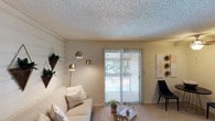 This single-floor residence offers a cohesive and thoughtfully designed living space that balances comfort with contemporary style. The living area serves as the heart of the home, featuring a textured ceiling that adds subtle dimension to the room. A large sliding glass door floods the space with natural light and provides a direct view of the outdoors, creating a seamless connection between the interior and exterior. Vertical blinds offer flexible light control and privacy as needed. The room is finished with plush carpeting underfoot, contributing to a warm and inviting atmosphere. A striking multi-arm brass arc floor lamp serves as both a functional and decorative focal point, complementing the neutral, light-toned furnishings that include a plush sofa adorned with layered throw pillows. An accent wall finished with white shiplap paneling adds architectural character and texture to the space. Decorative wall art and carefully placed plants introduce natural elements that enhance the overall ambiance. The layout is open and well-proportioned, allowing for comfortable movement and versatile furniture arrangement. Overall, the space presents a clean, modern aesthetic with warm accents, making it both functional and visually appealing.