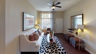 This single-story residence combines contemporary design with functional living across several well-defined spaces. The kitchen is a standout feature, showcasing dark espresso cabinetry, a mosaic tile backsplash, granite-style countertops, and a full suite of stainless steel appliances, including a refrigerator, electric range, microwave, and dishwasher. A built-in shelving nook adjacent to the kitchen doubles as a compact workspace or display area, adding versatility to the layout. Rich wood-style flooring runs through the kitchen and living areas, lending warmth and continuity. The living room is bright and welcoming, featuring a ceiling fan, large windows dressed with sheer paneled curtains, and natural light that enhances the neutral-toned walls. The bedroom offers plush carpeting, a ceiling fan, generous natural light from a well-proportioned window, and ample space for furnishings. The bathroom is cleanly appointed with a single vanity featuring granite-style countertops, a large mirror, and a full shower-tub combination. Overall, the space presents a cohesive, modern aesthetic with warm finishes throughout.