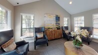 Located at 6100 Barrowood Dr in Raleigh, NC, this single-floor space offers a polished and functional interior designed to impress and accommodate. The open layout is anchored by dramatic vaulted ceilings that lend an airy, expansive feel throughout. Neutral warm-gray walls and patterned carpet tile flooring create a cohesive, professional aesthetic that flows seamlessly from one area to the next.

The lounge area is thoughtfully appointed with a pair of navy blue leather wingback chairs accented with decorative throw pillows, arranged around a round wooden coffee table. A richly textured wooden sideboard serves as a focal point, topped with curated décor and anchored by a large, striking abstract painting in navy, gold, and white tones. Multiple windows dressed with white horizontal blinds flood the space with natural light while offering views of the surrounding greenery.

Adjacent to the lounge, a dedicated workstation area features a modern desk with a computer and printer setup, ideal for business use. A wall-mounted television adds to the functionality of the space. French glass-panel doors connect the areas and allow light to pass freely, reinforcing the open, bright character of the interior. An exterior glass door provides direct access to the outdoors, further enhancing the sense of connectivity and light throughout this well-designed space.