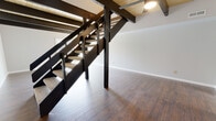This two-floor space combines modern design elements with functional living areas. The main level immediately impresses with its distinctive wood-planked ceiling adorned with bold, dark exposed beams that run throughout the open living area. A dramatic black open-riser staircase serves as a striking architectural focal point, connecting both floors while allowing light to flow freely through the space. Rich, dark hardwood flooring anchors the main level, contributing warmth and contrast against the crisp white walls.

The kitchen is thoughtfully designed in a galley layout, featuring white shaker-style cabinetry with matte black hardware, light countertops, a classic tile backsplash, and wood-accented surfaces. A window above the sink frames a lush, wooded outdoor view, filling the space with natural light.

The upper level transitions to soft, neutral carpeting throughout the carpeted bedrooms, each offering windows that bring in natural light and views of the surrounding greenery. Built-in shelving adds practical storage to the bedroom areas. The bathroom is finished with a deep navy vanity topped with a light stone-style countertop, dark hardwood-style flooring, and a full tub-shower combination, delivering a clean and polished aesthetic throughout.