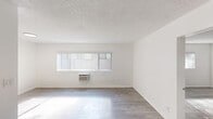 This single-floor unit offers a clean, modern aesthetic defined by a crisp white palette, light wood-style flooring, and thoughtfully updated finishes throughout. The open living area is bathed in natural light through a wide horizontal window fitted with vertical blinds, and features painted brick-textured walls that add subtle depth to the otherwise minimal space. A wall-mounted air conditioning unit provides climate control, while the textured ceiling adds a classic residential character.

The galley-style kitchen stands out as a striking design feature, showcasing deep navy blue shaker cabinetry paired with brushed gold hardware and white quartz countertops with elegant veining. Stainless steel appliances — including a refrigerator, microwave, and electric range — complete this well-appointed cooking space, which also includes a stainless undermount sink and tile flooring.

The bedroom offers a generous, open layout with ample wall space and access to a mirrored sliding closet, providing practical storage. Consistent light wood-style flooring flows throughout.

The bathroom is finished in a clean, all-white scheme featuring a quartz vanity top, a large frameless mirror, brushed nickel fixtures, and a full-size soaking tub with shower combo, creating a calm and functional space.