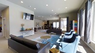 This single-floor space offers a thoughtfully designed open-concept layout that seamlessly blends comfort and functionality. The main lounge area is anchored by contemporary furnishings, including sleek leather sofas and modern accent chairs arranged around wooden coffee tables atop vibrant blue area rugs, creating a welcoming and visually dynamic environment. Recessed lighting throughout the ceiling provides consistent, warm illumination that complements the natural light streaming in through large windows dressed with floor-length curtains. A wall-mounted flat-screen television adds an entertainment focal point to the lounge, while a colorful vending machine tucked in the corner offers added convenience. Toward the back of the space, a kitchenette equipped with appliances and ample counter space provides a practical area for food preparation and casual dining. A foosball table is also visible, adding a recreational element that enhances the social atmosphere of the space. The neutral-toned walls and light-colored flooring create a clean, airy backdrop that ties together the modern décor. Overall, the space exudes a contemporary, community-oriented ambiance designed to encourage gathering, relaxation, and social interaction.