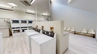 Located at 4400 Montgomery Blvd NE in Albuquerque, New Mexico, this shared laundry facility offers a clean, efficiently organized single-floor space designed to accommodate multiple users simultaneously. The interior is finished in a bright, neutral palette with light wood-look flooring and white walls that contribute to a fresh, welcoming atmosphere.

The facility is equipped with multiple top-load and front-load washing machines as well as stacked dryer units, providing ample laundry capacity. Generous built-in folding counters line the walls, offering dedicated surfaces for sorting and folding laundry with ease. A utility sink and additional workspace are also available for convenience.

The space features a distinct waiting area with molded chair seating beneath notably high vaulted ceilings, adding an unexpected sense of openness and volume to the room. A skylight overhead allows natural light to filter in, brightening the space considerably. Overhead linear fluorescent and pendant lighting ensures consistent illumination throughout.

Safety is addressed with a prominently mounted fire extinguisher, and HVAC equipment is visibly integrated for climate control. A partial dividing wall creates a natural separation between the active laundry area and the waiting zone, helping to organize the flow of the space. Overall, the facility is well-maintained and thoughtfully laid out for practical, communal use.