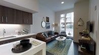 This single-floor space showcases a thoughtfully designed open-concept layout that seamlessly connects the kitchen and living areas. The kitchen is outfitted with dark wood cabinetry, a white quartz countertop, and a geometric mosaic tile backsplash, lending a refined, modern aesthetic. A white kitchen island provides additional prep and dining space, complemented by coordinating white lower cabinetry.

The living area is anchored by a rich brown leather sofa adorned with teal and patterned accent pillows, paired with a classic gray accent chair. A marble-topped coffee table with a gold base sits atop a blue vintage-style area rug, adding warmth and character to the space. Wall décor, including a round mirror and framed artwork, adds personality to the clean white walls.

A standout feature is the floor-to-ceiling window at the far end of the living area, which floods the space with natural light and offers street-level urban views. Dark hardwood flooring runs throughout, providing a grounding contrast to the bright walls and light furnishings. The overall ambiance is contemporary and inviting, balancing clean lines with warm, stylish accents.