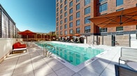 Located at 42 Broad St W in Mount Vernon, NY, this outdoor amenity deck offers a resort-inspired retreat set against the backdrop of a striking multi-story red brick building. The centerpiece of the space is a generously sized rectangular swimming pool finished with vibrant aqua mosaic tiles, giving the water a distinctive turquoise clarity. The pool transitions from a shallow entry to a deeper swimming area, accommodating a range of recreational uses.

Surrounding the pool is a well-appointed deck finished in light-toned large-format pavers, providing ample room for lounging and relaxation. Rows of contemporary chaise lounge chairs are positioned along the poolside, each shaded by bold orange umbrellas that add a lively pop of color to the environment. Additional seating vignettes with low-profile sofas and accent tables are tucked near warm wood-slatted privacy screens, creating more intimate outdoor living areas.

Architectural interest is added by a wood-beamed pergola structure on one end of the deck, offering filtered shade and a refined aesthetic. The warm, horizontal wood paneling along the building facade facing the pool provides a natural contrast to the surrounding brick architecture. The entire space is open to the sky, flooded with natural sunlight, making it a vibrant and welcoming outdoor destination for residents and guests alike.