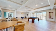 Located at 500 N Henry in Williamsburg, Virginia, this well-appointed amenity space spans a single floor and offers a thoughtfully designed layout ideal for gathering and recreation. The main game and lounge area is the centerpiece of the space, showcasing elegant coffered ceilings with beadboard detailing, recessed lighting, and wide-plank light hardwood flooring that flows throughout. A full-size pool table anchors the room, accompanied by stylish lounge seating and a dedicated round-table workspace with swivel chairs. Large French doors open to an outdoor balcony, flooding the interior with natural light and providing open views of the surrounding landscape. A separate dining area features bistro-style high-top tables and chairs beneath another coffered, beadboard ceiling, with oversized windows that continue the bright, airy aesthetic. Contemporary artwork adorns the crisp white walls throughout, adding color and character. The kitchen is finished with white shaker cabinetry accented by gold hardware, quartz countertops, a stainless steel refrigerator, microwave, and a wall-mounted flat-screen television. Tile backsplash detailing and an island sink complete the space. The overall ambiance is clean, modern, and inviting, blending classic architectural elements with contemporary furnishings.