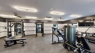 Located at 968 Bonnel Ct in Union, New Jersey, this fitness center offers a well-appointed exercise room designed to support a comprehensive workout experience. The space features a modern aesthetic, with textured wall paneling in contrasting dark and light tones, durable speckled rubber flooring, and a clean white ceiling outfitted with sleek oval recessed lighting fixtures and a ceiling fan for air circulation. A dedicated HVAC cassette unit ensures climate control throughout.

The exercise room is fully equipped with a robust selection of Matrix-brand machines, including a functional trainer, cable systems, a stair climber, and multiple selectorized strength stations. A row of treadmills lines one wall, accompanied by a mounted flat-screen display for entertainment during cardio sessions. Free weight stations are well-represented, with a full dumbbell rack, adjustable benches, and a power rack for resistance training.

Large floor-to-ceiling windows and glass partition walls on one side of the room allow ample natural light to filter in and provide views into adjacent common areas. Wall-mounted mirrors further enhance the sense of openness. The overall ambiance is clean, professional, and purposefully designed to deliver a high-quality fitness facility.