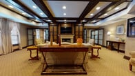 This single-floor space exudes a refined, club-like atmosphere defined by rich dark wood millwork, coffered ceilings with recessed lighting, and warm neutral carpeting throughout. The central lounge area serves as the heart of the space, anchored by a traditional white fireplace framed by dark wood built-in cabinetry and a wall-mounted television above. Ornate decorative columns flank the entertainment wall, adding architectural character. Elegant furnishings — including upholstered sofas, carved wooden accent chairs, and cherry-toned side tables — are arranged for comfortable conversation and relaxation.

An adjacent seating area features a grand piano alongside multiple armchairs and sofas, bathed in natural light from large windows dressed with floor-length drapes. Frosted glass French doors provide transitions between spaces while maintaining an open, cohesive feel. A separate dining area with a round table and high-back chairs is visible through a connecting corridor lined with windows and white shutters.

Additional rooms accessible via frosted double doors offer flexible use, decorated with patterned wallpaper and framed artwork. Throughout, the consistent use of dark wood trim, neutral tones, and classic furnishings creates a warm, sophisticated ambiance.