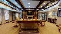This single-floor space exudes a refined, club-like atmosphere defined by rich dark wood millwork, coffered ceilings with recessed lighting, and warm neutral carpeting throughout. The central lounge area serves as the heart of the space, anchored by a traditional white fireplace framed by dark wood built-in cabinetry and a wall-mounted television above. Ornate decorative columns flank the entertainment wall, adding architectural character. Elegant furnishings — including upholstered sofas, carved wooden accent chairs, and cherry-toned side tables — are arranged for comfortable conversation and relaxation.

An adjacent seating area features a grand piano alongside multiple armchairs and sofas, bathed in natural light from large windows dressed with floor-length drapes. Frosted glass French doors provide transitions between spaces while maintaining an open, cohesive feel. A separate dining area with a round table and high-back chairs is visible through a connecting corridor lined with windows and white shutters.

Additional rooms accessible via frosted double doors offer flexible use, decorated with patterned wallpaper and framed artwork. Throughout, the consistent use of dark wood trim, neutral tones, and classic furnishings creates a warm, sophisticated ambiance.