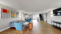 This thoughtfully designed, single-floor communal space offers a generous open-plan layout finished with warm hardwood flooring throughout. The interior is bathed in natural light from large windows along the perimeter, providing views of the surrounding neighborhood and creating a bright, welcoming atmosphere.

The space is organized into distinct functional zones. A central lounge area features a grey sofa, bold blue accent chairs, and a sleek coffee table with tufted orange ottomans, striking a balance between comfort and contemporary style. Adjacent to this, a dedicated dining and breakout nook is anchored by a marble-topped round table with teal chairs, set against a vibrant geometric mural rendered in blue, coral, and lavender tones — a clear artistic focal point of the room.

A separate seating alcove near the entrance offers additional flexible gathering options alongside a wall-mounted television and a slim console table. A credenza with a coffee station and ample counter space serves a practical function in one corner, enhanced by a nearby window that fills the area with daylight.

A staircase leads to an upper level, hinting at additional connected space. Wall sconces complement the recessed and track lighting, ensuring the space is well-lit at all times. The clean white walls, crown molding details, and curated décor come together to create a polished yet inviting communal environment.
