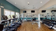Located on N Ben Wilson Street in Victoria, Texas, this fitness center is a well-equipped, single-floor facility designed to support a comprehensive workout experience. The space features a distinctive two-tone color scheme, with deep teal accents along the lower walls contrasting against neutral beige upper walls and white ceilings, creating an energetic yet focused atmosphere.

The main workout area is generously sized and thoughtfully organized into distinct zones. Cardio equipment — including treadmills, elliptical trainers, stationary bikes, and a rowing machine — lines the perimeter, with large windows offering views of the outdoor surroundings and filling the space with natural light. Ceiling fans and recessed lighting ensure year-round comfort and visibility.

The strength training section is centrally positioned and features a power rack, adjustable benches, a full dumbbell rack with a wide range of weights, and a cable/weight machine. Wall-mounted mirrors span a significant portion of the facility, visually expanding the space and providing form-check visibility throughout.

A dedicated functional training area includes stability balls, medicine balls, and an adjustable workout bench, ideal for stretching and dynamic exercises. Multiple wall-mounted flat-screen televisions are positioned throughout for entertainment during workouts. Durable hardwood-style flooring ties the entire space together, offering both practical durability and a polished aesthetic.