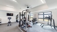This fitness center in Huntsville, Alabama offers a generously sized, single-floor layout designed to accommodate a comprehensive range of workout activities. The space features soaring ceilings that enhance the open, airy feel of the interior, while large windows along the walls flood the room with natural light and provide views of the surrounding outdoor environment.

The facility is divided into clearly defined workout zones. The strength training area is anchored by a multi-functional cable and Smith machine system, accompanied by a full dumbbell rack with an adjustable bench, making it suitable for a wide variety of free weight and resistance exercises. A dedicated cardio section houses multiple treadmills, elliptical machines, a stair climber, and rowing machines, all positioned to take advantage of the natural light streaming through the windows.

Wall-mounted flat-screen televisions are strategically placed throughout the space to keep users engaged during workouts. Ceiling fans maintain air circulation across the room, contributing to a comfortable exercise environment. The floors are finished with durable rubber-composite flooring suited for high-traffic fitness use. A water fountain station is also available. The overall aesthetic is modern and clean, with crisp white walls and a well-organized layout.