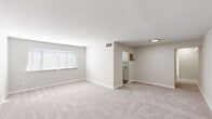 This single-floor unit is designed with a clean, neutral aesthetic, featuring light gray walls, white trim, and plush wall-to-wall carpeting across the living and bedroom areas. The spacious living room is a standout feature, offering an expansive open layout filled with natural light from a wide multi-pane window fitted with horizontal blinds. A pass-through opening connects the living area to the kitchen, enhancing the sense of openness between spaces.

The kitchen is efficiently laid out with white cabinetry, granite-look countertops, a stainless steel sink, and a gas range. Light wood-look flooring and a window above the cooking area keep the space bright and functional.

Multiple bedrooms share a consistent design language — neutral carpet, light walls, and individual windows that allow natural light to filter in. Each room is generously proportioned, providing comfortable living space. Ceiling-mounted light fixtures are present in the bedrooms.

The bathroom is finished in an all-white palette, featuring a tub-shower combination with white tile surround, a white vanity with brushed nickel fixtures, a framed mirror, and globe vanity lighting above. The overall unit presents a well-maintained, move-in ready environment with a cohesive, understated design throughout.