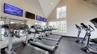 Located at 4890 Edwards Farm Rd in Hilliard, Ohio, this single-floor fitness center is a well-appointed facility designed to support a comprehensive workout experience. The space is defined by dramatic vaulted ceilings that rise to impressive heights, creating an open and airy atmosphere throughout. Large multi-pane windows line the walls, flooding the interior with natural light and offering views of the surrounding outdoor environment.

The fitness center is thoughtfully organized into distinct workout zones. A dedicated cardio area houses a row of treadmills and elliptical machines, each equipped with individual screens for entertainment during workouts. Multiple wall-mounted flat-screen televisions are positioned throughout for easy viewing. A full-length mirror along one wall enhances the sense of space in this section.

A separate free weights area features a well-stocked dumbbell and kettlebell rack, a weight bench, and stability balls, all positioned beneath bright windows. Rowing machines and stationary bikes round out the equipment selection, providing a full range of cardiovascular and strength training options.

The facility benefits from durable dark rubber flooring throughout, and floor-to-ceiling glass panel walls on one side allow sightlines across the full length of the space. Multiple entry and exit doors, including a large garage-style glass door, provide easy access and enhance ventilation and connectivity to the outdoors.