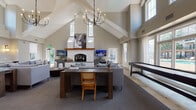 The clubhouse at Francis Way in Bloomfield, CT is a thoughtfully designed single-story community gathering space that balances comfort with contemporary style. The main lounge area is the architectural centerpiece, showcasing dramatic vaulted ceilings with exposed white beams that rise to impressive heights, flooding the space with natural light through an abundance of windows. A striking white stone fireplace with a warm wood mantel serves as the focal point, complemented by multiple flat-screen televisions and an elegant wrought-iron chandelier overhead. Generous seating arrangements include plush gray sofas arranged around coffee tables, creating inviting conversation areas. A large wooden table with cane-back chairs provides a flexible workspace or gathering spot. Additional amenities include a shuffleboard table and built-in desk nooks along windowed walls, ideal for quiet work or study. The kitchen area features rich dark wood cabinetry, a marble-topped island with bar seating, stainless steel appliances including a full-size refrigerator and dishwasher, and open navy floating shelves for display. Light hardwood flooring transitions between areas, while soft neutral tones throughout create a cohesive, welcoming ambiance suited for both casual socializing and organized community events.