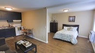 This single-floor unit offers an open-concept layout that efficiently combines living, sleeping, and dining functions within one cohesive space. Warm hardwood flooring extends throughout, providing a consistent and inviting foundation that ties the areas together visually.

A partial wall creates a subtle division between the sleeping area and the rest of the unit, offering a degree of privacy without fully closing off the space. The sleeping area accommodates a full-sized bed complemented by a tall dark-wood dresser and framed wall art, giving it a finished, comfortable feel.

Adjacent to the living area, a small dining nook features a compact bistro-style table and chairs, making practical use of the available space. A low-profile coffee table anchors the living area in the foreground.

The walls are finished in warm beige tones, contributing to the overall cozy ambiance. A textured ceiling adds subtle visual interest overhead, while flush-mounted lighting ensures the space remains well lit. The open layout and neutral palette make the unit feel airy and adaptable to a variety of furnishing styles.