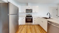 This single-floor residence is designed with a clean, modern aesthetic and a well-organized layout that maximizes functionality throughout. The open-concept living and kitchen area serves as the heart of the home, where warm-toned hardwood flooring extends seamlessly across the space. Recessed lighting illuminates the area, while multiple windows along one wall invite generous natural light and offer views of the surrounding exterior, complemented by a door leading to a private balcony.

The kitchen is thoughtfully appointed with white shaker-style cabinetry, marble-look countertops, and a central island with a built-in sink. Stainless steel appliances — including a refrigerator, electric range, and over-the-range microwave — provide a polished, contemporary feel. The island creates a natural divide between the kitchen and the expansive living area.

The bedrooms are carpeted for comfort, each featuring crisp white walls, ample closet space with double doors, and a quiet, neutral atmosphere. The bathroom continues the home's consistent design language with hardwood flooring, a white vanity, and black hardware accents. A dedicated laundry area includes a full-sized top-loading washer and dryer, adding everyday convenience to this well-appointed space.