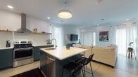 Located at 118 SE I St in Bentonville, Arkansas, this single-story space offers a thoughtfully designed open-concept layout that seamlessly connects the kitchen, dining, and living areas. The kitchen is a standout feature, showcasing bold teal cabinetry paired with crisp white upper cabinets and countertops, stainless steel appliances including a range with a statement hood vent, a built-in refrigerator, and a central island with a gold-toned faucet and bar seating. Open wood shelving adds a warm, functional touch. The living area is bright and inviting, benefiting from natural light streaming through large sliding glass doors dressed with vertical blinds and sheer curtains. A built-in niche with shelving and teal accent walls provides a stylish focal point. The bedroom is a serene retreat featuring direct outdoor access, sheer curtain-draped windows, and a built-in desk nook with open shelving — ideal as a workspace. The bathroom is clean and modern, with a floating white vanity, tiled tub-shower combination, and polished concrete floors that continue throughout the unit. A dedicated laundry area with a stacked washer and dryer, clothing rods, and storage shelving rounds out this well-appointed space.