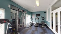 This fitness center is a compact yet comprehensively outfitted space designed to support a well-rounded workout routine. The room is finished with distinctive teal-painted walls and crisp white trim, creating an energizing and clean atmosphere. Striped, multi-toned carpet tiles cover the floor throughout, adding both durability and visual interest underfoot.

The space is equipped with a variety of cardio machines, including treadmills, elliptical trainers, and a stationary cycling bike, providing options for diverse aerobic workouts. A Hoist multi-function cable machine offers strength training capabilities, complemented by a set of kettlebells for free-weight exercises. A wall-mounted flat-screen television is positioned for easy viewing during workouts, and a full-length mirror along one wall enhances the sense of space while aiding in form and technique. Workout reference posters are displayed on the walls for additional guidance.

Natural light filters in through large windows fitted with horizontal blinds, brightening the interior during daytime hours. Recessed and overhead lighting ensures the space remains well-lit at all times. The room also features a clearly marked exit door, ensuring safe and convenient access. The overall layout is open and functional, making efficient use of the available space for fitness activities.