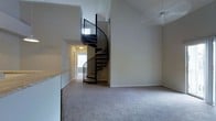 This space makes a strong architectural statement with its eye-catching black metal spiral staircase, which serves as a central focal point and rises toward notably high, vaulted ceilings that lend an impressive sense of vertical space and openness. The living area is finished with plush, neutral-toned carpeting that adds warmth and comfort underfoot, while the surrounding walls are painted in a clean, soft beige palette that enhances the bright, airy feel of the interior.

The open layout connects the living area to an adjacent hallway corridor, which is illuminated by natural light filtering in from a window visible at the far end, contributing to a well-lit and welcoming atmosphere. A partial view of what appears to be a kitchen counter with light granite or stone-finish surfaces is visible to the left, suggesting a practical and connected layout between the kitchen and living spaces.

The vaulted ceiling design amplifies the sense of scale throughout, making the main living area feel generously proportioned. Recessed lighting and smoke detection fixtures are integrated into the ceiling. Overall, the space balances functional design with a distinctive architectural element in the spiral staircase, creating a memorable and characterful interior.