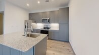 This thoughtfully designed 2-bedroom, 2-bathroom residence is laid out on a single floor, offering a functional and cohesive living experience. Light wood-grain flooring runs throughout the space, tying together each room with a clean, contemporary aesthetic.

The kitchen serves as a central highlight, featuring floor-to-ceiling gray shaker-style cabinetry with brushed nickel hardware, a white hexagonal tile backsplash, and stainless steel appliances including a refrigerator, range, and microwave. A kitchen island with a granite countertop and undermount sink provides additional prep space and casual seating potential. Pendant lighting and recessed fixtures illuminate the area effectively.

The open living area adjoins the kitchen and benefits from natural light via large gridded windows, a ceiling fan, and direct access to an outdoor balcony through a glass-panel door.

The spacious primary bedroom features multiple windows for ample natural light, a ceiling fan, and access to a walk-in closet with built-in shelving. The second bedroom mirrors similar finishes and also enjoys natural light through gridded windows.

Both bathrooms are finished consistently, each offering a gray shaker vanity with granite countertops, a framed mirror, modern light fixtures, floating shelves, and tile flooring. One bathroom includes a soaking tub with tiled surround, while the other features a glass-enclosed shower.