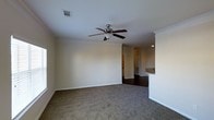 This single-floor residence offers a comfortable and functional layout designed for everyday living. The main living area is finished with plush carpeting and features elegant crown molding that frames the ceiling, adding a refined architectural touch throughout the space. A ceiling fan with a built-in light fixture is centrally mounted, providing both air circulation and ambient lighting. The neutral wall tones create a clean, versatile backdrop that complements a variety of interior styles. The living space transitions seamlessly into the kitchen area, which is distinguished by dark hardwood-style flooring that contrasts with the lighter tones of the living room. A raised breakfast bar serves as a natural dividing element between the two spaces, making it ideal for casual dining or entertaining. The kitchen area is bright and functional, with cabinetry visible in the background. Ample ventilation is provided through ceiling-mounted HVAC vents, ensuring year-round comfort. The open-concept flow between the living and kitchen areas enhances the sense of space, making the home feel connected and inviting. The overall ambiance is clean, move-in ready, and well-suited for a range of living arrangements.
