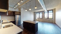 This single-floor urban loft showcases a compelling blend of industrial character and modern finishes throughout its open layout. The living and kitchen areas flow seamlessly together, anchored by striking architectural details including exposed silver ductwork, painted corrugated ceilings, and prominent decorative columns that speak to the building's converted heritage.

The kitchen is a standout feature, offering rich dark-walnut cabinetry, light-toned countertops, and a full suite of stainless steel appliances including a refrigerator, range, and built-in microwave. A central island provides additional prep and seating space, while pendant lighting overhead adds warmth and ambiance. Track lighting runs along the ceiling, providing flexible illumination across the open-plan space.

The generously proportioned living area is bathed in natural light from large, black-framed windows that offer views of the surrounding cityscape and skyline. Light hardwood flooring runs throughout, complementing the bright, airy atmosphere.

The bathroom is finished with a clean, contemporary aesthetic, featuring large-format tile flooring, a vanity with dark countertop and under-mount sink, and a walk-in shower enclosed by sliding glass doors with tiled surround. The overall ambiance of this loft is one of industrial elegance — spacious, light-filled, and thoughtfully designed.
