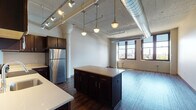 This single-floor urban loft showcases a compelling blend of industrial character and modern finishes throughout its open layout. The living and kitchen areas flow seamlessly together, anchored by striking architectural details including exposed silver ductwork, painted corrugated ceilings, and prominent decorative columns that speak to the building's converted heritage.

The kitchen is a standout feature, offering rich dark-walnut cabinetry, light-toned countertops, and a full suite of stainless steel appliances including a refrigerator, range, and built-in microwave. A central island provides additional prep and seating space, while pendant lighting overhead adds warmth and ambiance. Track lighting runs along the ceiling, providing flexible illumination across the open-plan space.

The generously proportioned living area is bathed in natural light from large, black-framed windows that offer views of the surrounding cityscape and skyline. Light hardwood flooring runs throughout, complementing the bright, airy atmosphere.

The bathroom is finished with a clean, contemporary aesthetic, featuring large-format tile flooring, a vanity with dark countertop and under-mount sink, and a walk-in shower enclosed by sliding glass doors with tiled surround. The overall ambiance of this loft is one of industrial elegance — spacious, light-filled, and thoughtfully designed.