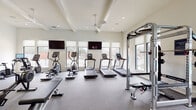 Located at 3612 Beverly Cove Way in Wilmington, North Carolina, this single-story fitness center is thoughtfully designed to accommodate a comprehensive range of workout needs. The facility is divided into distinct functional zones, each finished with attention to both aesthetics and practicality.

The main cardio area features multiple treadmills, ellipticals, and cycling machines arranged along a wall of large windows, flooding the space with natural light and offering outdoor views. Ceiling fans and recessed lighting maintain a comfortable environment, while wall-mounted televisions provide entertainment during workouts. Durable rubber flooring throughout this area ensures safety and sound absorption.

The strength training zone includes a multi-functional cable rack, free weights, benches, a rowing machine, and a power rack, all set beneath coffered ceilings with additional ceiling fan coverage. Full-length mirrors line the walls, enhancing the sense of openness.

A separate studio room, enclosed by sleek black-framed glass partition doors, offers light hardwood flooring, soaring ceilings, decorative pendant lighting, and floor-to-ceiling mirrors—ideal for stretching, yoga, or functional training with equipment such as stability balls and adjustable benches.

The facility also includes an accessible unisex restroom, distinguished by a striking geometric-patterned tile floor and clean white beadboard wainscoting.