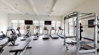 Located at 3612 Beverly Cove Way in Wilmington, North Carolina, this single-story fitness center is thoughtfully designed to accommodate a comprehensive range of workout needs. The facility is divided into distinct functional zones, each finished with attention to both aesthetics and practicality.

The main cardio area features multiple treadmills, ellipticals, and cycling machines arranged along a wall of large windows, flooding the space with natural light and offering outdoor views. Ceiling fans and recessed lighting maintain a comfortable environment, while wall-mounted televisions provide entertainment during workouts. Durable rubber flooring throughout this area ensures safety and sound absorption.

The strength training zone includes a multi-functional cable rack, free weights, benches, a rowing machine, and a power rack, all set beneath coffered ceilings with additional ceiling fan coverage. Full-length mirrors line the walls, enhancing the sense of openness.

A separate studio room, enclosed by sleek black-framed glass partition doors, offers light hardwood flooring, soaring ceilings, decorative pendant lighting, and floor-to-ceiling mirrors—ideal for stretching, yoga, or functional training with equipment such as stability balls and adjustable benches.

The facility also includes an accessible unisex restroom, distinguished by a striking geometric-patterned tile floor and clean white beadboard wainscoting.