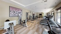 This modern fitness center is thoughtfully designed to support a comprehensive workout experience across two distinct areas. The main exercise floor features a generous selection of cardio equipment, including multiple treadmills, elliptical machines, and stationary bikes, all positioned alongside large floor-to-ceiling windows that flood the space with natural light and offer open views of the surrounding streetscape. A row of open shelving units provides convenient storage, while wall-mounted flat-screen televisions keep members engaged during their sessions. Recessed ceiling lighting and a tray ceiling detail lend a polished, upscale ambiance to the main floor. Neutral-toned walls and a distinctive checkered carpet tile flooring in warm beige tones unify the space with a clean, contemporary aesthetic. A dedicated functional training studio adjoins the main area, finished with the same patterned flooring and accented by soft yellow-green walls. This studio is equipped with a wall-mounted climbing frame, TRX suspension training straps, and a large mirrored wall that enhances the sense of space and supports form monitoring. An additional flat-screen television is mounted within the studio for guided workout viewing. Together, the two areas create a versatile and well-appointed fitness environment suited for a wide range of training styles.