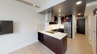 This well-appointed one-bedroom, one-bathroom unit is designed with a modern aesthetic, offering a thoughtful layout across a single floor. The kitchen is a standout feature, showcasing rich dark-wood cabinetry with frosted glass upper panels, light stone countertops, and a full suite of stainless steel appliances, including a refrigerator, dishwasher, and cooktop. A peninsula with a matching countertop extends into the living area, providing additional prep space and a natural division between the kitchen and living spaces. A wall-mounted range hood adds an industrial-chic touch, while track lighting illuminates the area effectively.

The living area is bright and open, featuring light wood-style flooring that runs throughout the unit. Two large windows flood the space with natural light and frame city views, enhancing the airy atmosphere. The clean white walls and neutral tones create a calm, versatile backdrop.

The bathroom is clean and functional, featuring light wood-style tile flooring, a dark vanity with a stone-topped sink, a full tub with sliding glass shower doors, and ample towel storage. The overall design of the unit balances contemporary style with practical functionality.