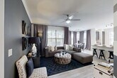 This well-appointed 2-bedroom, 4-bathroom single-story residence offers a thoughtfully designed floor plan that maximizes comfort and functionality. The open-concept living and dining area is bathed in natural light from large windows dressed with elegant floor-to-ceiling drapes, creating a bright and inviting atmosphere. The living room features stylish touches including a bold dark accent wall adorned with a gallery of gold-framed mirrors, wood-look flooring, and a herringbone-patterned area rug. The kitchen is a standout, equipped with rich dark cabinetry, granite countertops, a subway tile backsplash, stainless steel appliances, and a central island — all illuminated by a large window with outdoor views. The generously sized primary bedroom features plush carpeting, decorative wallpaper, a ceiling fan, and ample natural light. Bathrooms are finished with granite vanities, dark cabinetry, and clean white tile tub surrounds. Additional highlights include a dedicated laundry room with full-size washer and dryer, a closet, a utility room, and a hallway connecting the well-organized layout. The overall aesthetic is modern and sophisticated, with a cohesive neutral and dark-toned palette throughout.