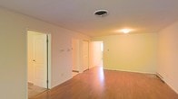 This single-floor space presents a clean, straightforward layout designed for practical living. The main open area is finished with warm-toned hardwood flooring that reflects light and adds a sense of warmth throughout the room. The walls are painted in a soft, neutral cream hue, creating a bright and welcoming atmosphere that complements the flooring.

The space features multiple doorways leading to additional rooms, suggesting a well-connected floor plan. A central doorway with a paneled white door provides access to what appears to be a brighter adjoining room, possibly receiving natural light from a nearby window. Additional doorways on either side of the main area offer further connectivity throughout the unit.

Ceiling-mounted HVAC vents ensure climate control throughout, while flush-mounted ceiling lighting provides functional illumination. White baseboards frame the perimeter of the room, adding a clean, finished look to the overall space. The neutral palette and hardwood flooring throughout create a cohesive aesthetic that is both versatile and easy to personalize, making it well-suited for a variety of furnishing styles and uses.