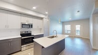 Located at 2700 W Cobalt Dr in Meridian, Idaho, this single-floor unit offers a clean, modern design with thoughtful finishes throughout. The open-concept kitchen serves as the heart of the space, showcasing a central island with a white quartz countertop and undermount sink, complemented by sleek pendant lighting. Tall white cabinetry lines the walls, providing ample storage, while a full suite of stainless steel appliances — including a refrigerator, electric range, and built-in microwave — adds a polished, contemporary feel. Light wood-look flooring runs through the main living areas, lending warmth and continuity to the open layout. Natural light flows in through multiple windows fitted with horizontal blinds, brightening both the living area and the entryway. The entry door features a sidelight panel, welcoming additional daylight into the foyer. Bedroom areas are finished with plush carpet and include dedicated closet space with built-in rods and recessed lighting. The bathroom features a full soaking tub and shower combination with clean white subway tile surrounds and chrome fixtures. A dedicated laundry area houses a full-size top-loading washer and dryer, adding everyday convenience. The overall aesthetic is bright, neutral, and move-in ready.