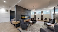 Located at 1100 Avenue du Docteur-Penfield in Montréal, this sophisticated lounge offers a thoughtfully designed communal space spread across a single floor. The interior is anchored by a dramatic floor-to-ceiling concrete-clad fireplace wall, which serves as the focal point of the main seating area. Plush tufted armchairs, a grey sofa with accent cushions, and a live-edge wood coffee table on hairpin legs create an inviting and contemporary gathering space. The herringbone-patterned hardwood flooring flows throughout, lending warmth and visual continuity to the entire lounge. A media area features a large wall-mounted television set against a dark accent wall, complemented by a sleek wood media console and a statement egg-style swivel chair. Slim, horizontal windows along one wall invite natural light while maintaining privacy. Dark wood slatted partitions elegantly divide the space, leading to a dining and kitchenette area outfitted with a white quartz-topped bar counter, pendant lighting, and vibrant blue seating. Wall sconces, globe pendant fixtures, and recessed lighting work together to create a warm, layered ambiance throughout. A large-format cityscape mural near the entrance adds a bold urban character to this polished and functional lounge.