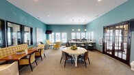This single-floor community space is defined by its cohesive teal-painted walls and neutral carpet flooring, creating a calm and inviting atmosphere throughout. The main gathering area is thoughtfully divided into distinct functional zones that flow seamlessly into one another. A central lounge area features a mix of yellow upholstered armchairs and a light blue accent chair arranged around a rustic wood coffee table, complemented by a wall-mounted flat-screen television and abstract artwork. Adjacent to this is a dining area with a round white pedestal table and dark wood chairs, as well as a separate rectangular dining table with bench-style tufted seating and slipcover chairs. A row of dark-framed interior windows lines one wall, offering views into a fitness center beyond, effectively connecting the spaces visually. The kitchenette area is well-appointed with dark wood cabinetry, light granite countertops, a coffee station, microwave, and sink, bathed in natural light from multiple large windows with white blinds. Recessed lighting and pendant fixtures illuminate the space throughout, while high ceilings contribute to an open, airy feel. The overall design balances comfort and functionality with a consistent, polished aesthetic.
